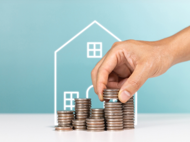 a drawing of a house behind stacks of coins and a human hand