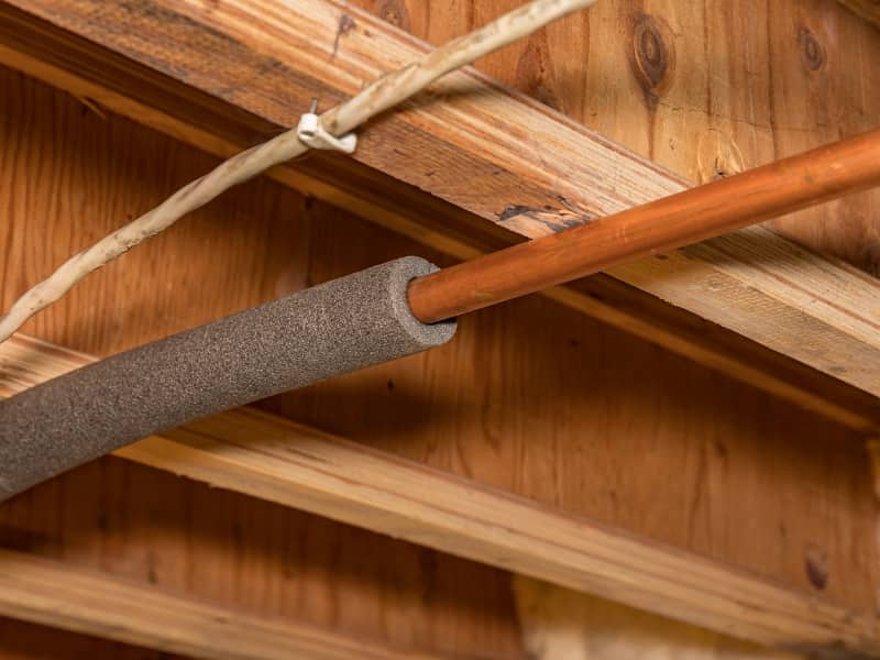 Foam insulation being fitted over a copper pipe beneath a wooden ceiling joist
