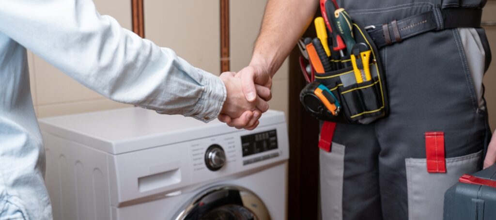 Customer shaking hands with a plumber in uniform, standing next to a washing machine with visible tools in a belt pouch.