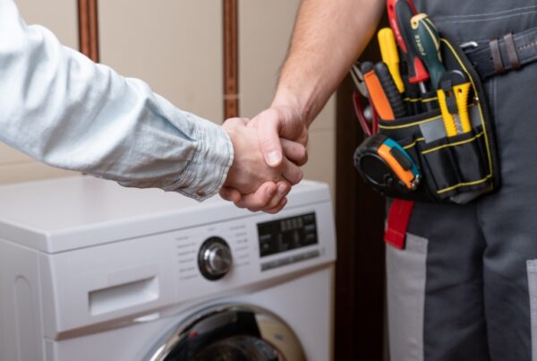 Customer shaking hands with a plumber in uniform, standing next to a washing machine with visible tools in a belt pouch.
